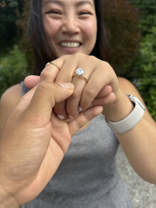 An engaged couple stands together, the woman smiling and holding out her hand to display an engagement ring with a large diamond. The man's hand holds her hand tenderly, highlighted against a backdrop of lush greenery. She is wearing a gray sleeveless outfit and a white smartwatch on her wrist.